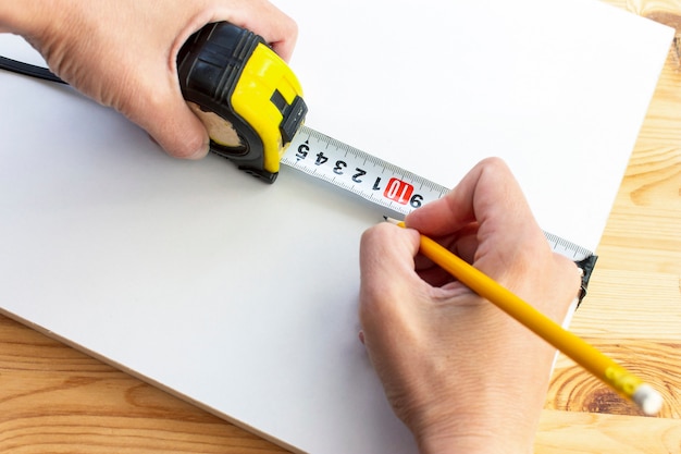 Cropped hands of a carpenter marking white wooden panel with yellow pencil and tape ruler