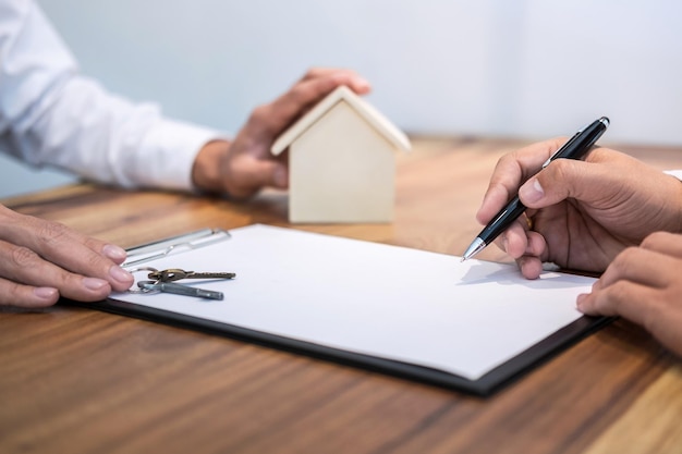 Cropped hands of client signing contract paper by businessman with house key on desk