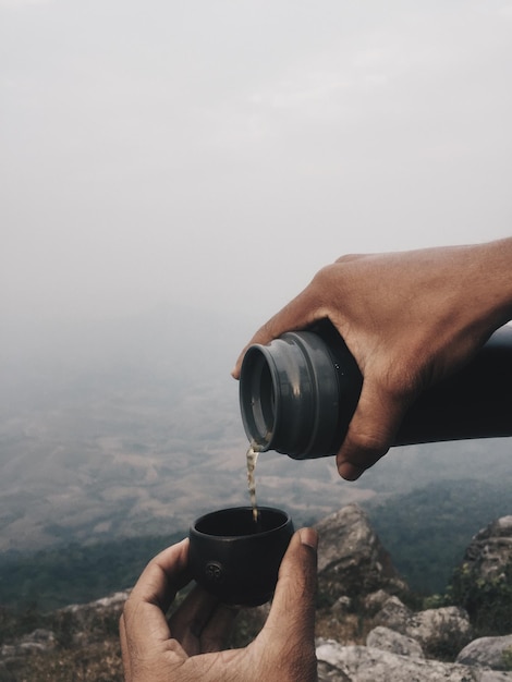 霧の天候で風景に照らして ⁇ 蓋に飲み物を注ぐ手を切った