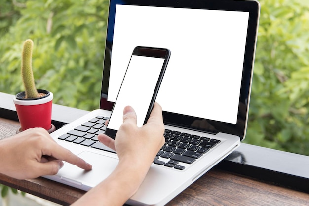 cropped hands of woman using mobile phone and laptop by window at table