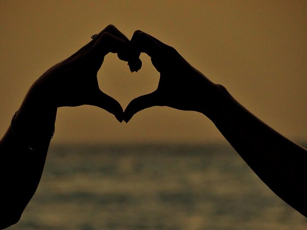 Photo cropped hands of women making heart shape against sky during sunset