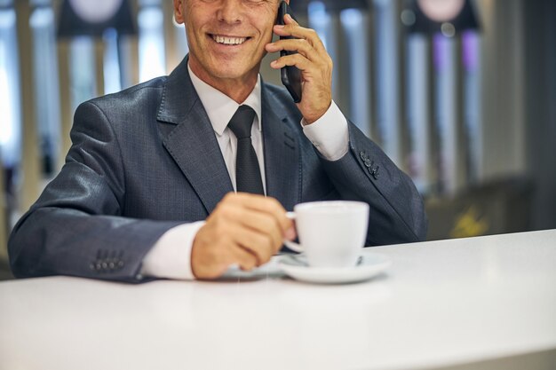 Testa tagliata di un maschio elegante e allegro che si gode una tazza di caffè mentre parla al cellulare