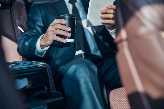 Cropped head of elegant man in suit and tie sitting in back\
with cup of hot drink and touchpad during transfer