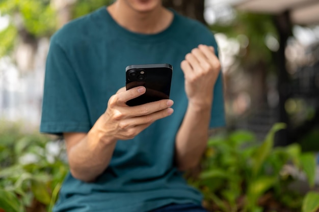 A cropped image of an Asian man in casual wear sitting in a city park and using his smartphone