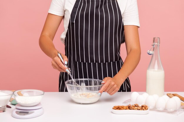 Cropped image of baker in black striped apron mixing dough with corolla making bakery homemade pastry confectioner preparing cake Indoor studio shot isolated on pink background