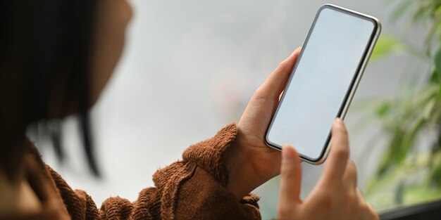 座っていると快適なリビングルームで彼女の休日にリラックスしながら白い空白の画面のスマートフォンを手に持った茶色の綿のセーターで美しい女性のトリミングされた画像。