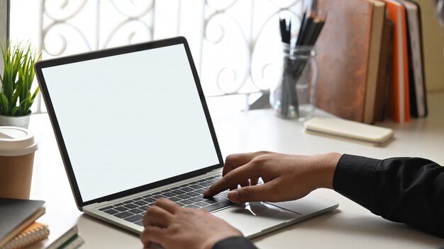 Cropped image of businessman hands typing on computer laptop with white blank screen that putting on orderly working desk that surrounded with books, pencil holder, coffee cup and potted plant.