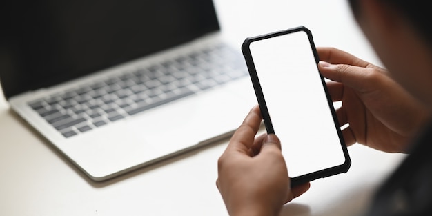Cropped image of businessman's hands holding a cropped black smartphone with white blank screen putting on modern white table in front computer laptop.