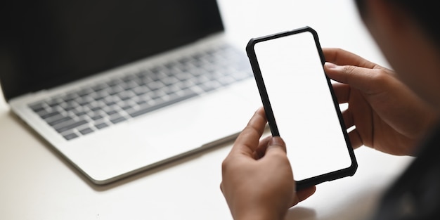 前面のコンピューターのラップトップのモダンな白いテーブルに置く白い空白の画面でトリミングされた黒いスマートフォンを持っているビジネスマンの手の画像をトリミングしました。
