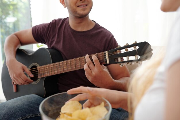 ポテトチップスの大きなボウルで彼のガールフレンドのためにギターを弾く陽気な若い男のトリミングされた画像