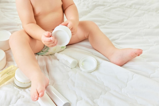 Cropped image of child legs toddler sitting in bed with body moisturizing creams lotions skincare