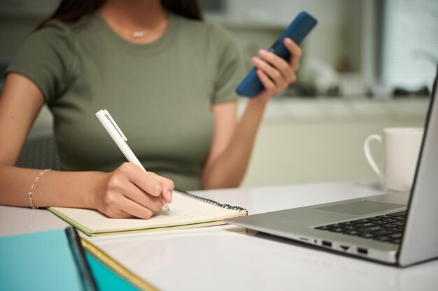 Cropped image of college student listening to voice message and taking notes in textbook