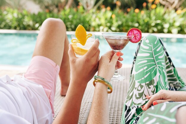 Photo cropped image of couple drinking cocktails when relaxing on chaise-lounges by swimming pool