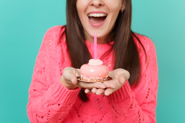 Cropped image of excited young woman in knitted pink sweater holding in hands cake with candle isolated on blue turquoise wall background studio portrait. People lifestyle concept. Mock up copy space.