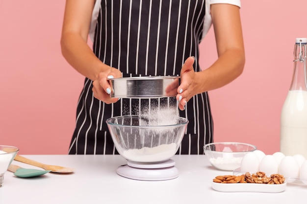 Обрезанное изображение женщины-кондитера просеивает муку для приготовления домашней выпечки, дама в фартуке держит сито в руках, окруженная ингредиентами для выпечки. Крытая студия снята на розовом фоне