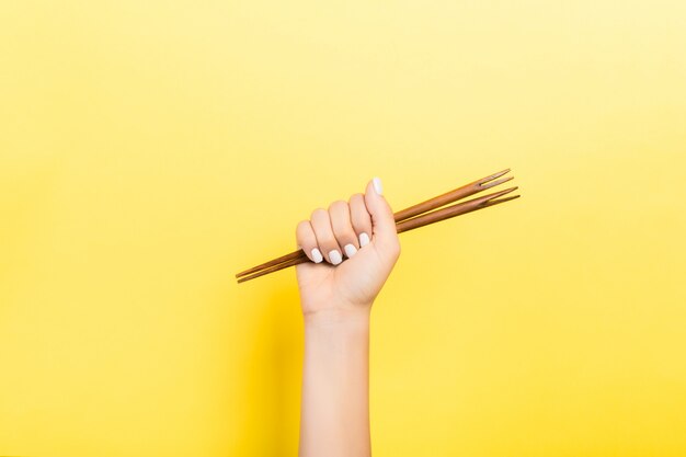 Cropped image of female hand holding chopsticks in fist on yellow background. Asian food concept with copy space