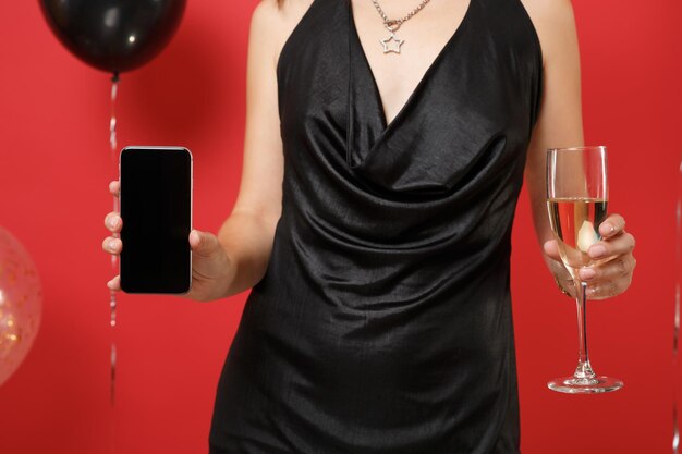 Immagine ritagliata della ragazza in abito nero che tiene in mano un bicchiere di champagne, telefono cellulare con schermo vuoto nero vuoto su mongolfiere di sfondo rosso brillante. felice anno nuovo compleanno mockup festa concetto.