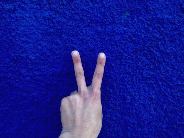 Photo cropped image of hand gesturing peace sign against blue wall