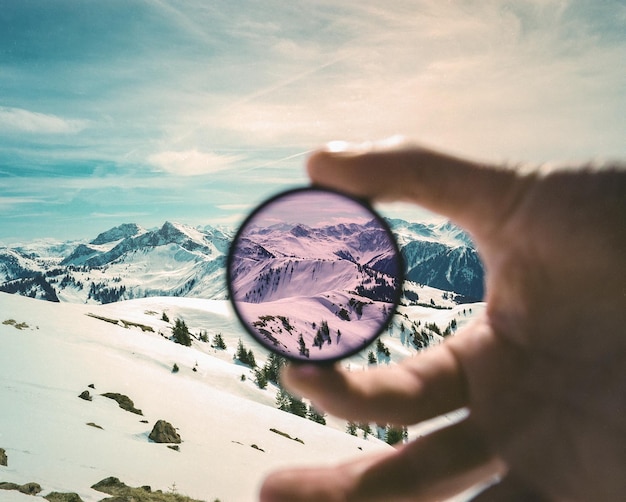 Cropped image of hand holding circular glass against snowcapped mountains