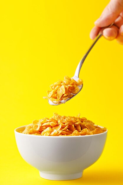 Cropped image of hand holding cornflakes in spoon over bowl against yellow background