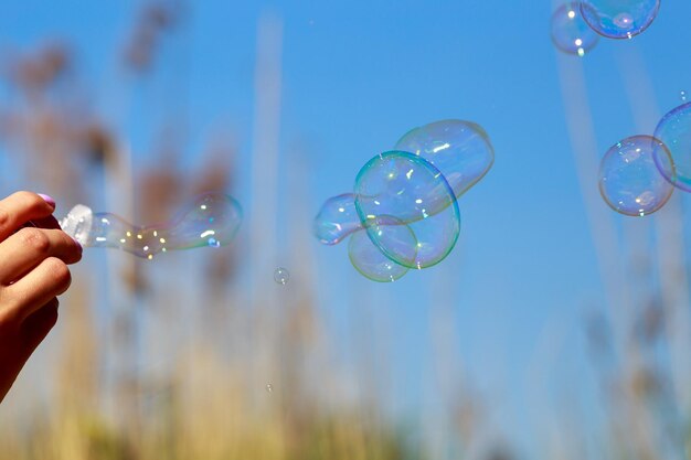 Cropped image of hand holding wand with bubbles flying against blue sky