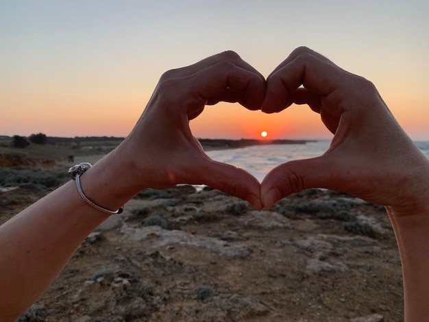 Cropped image of hands making heart shape against sky during sunset