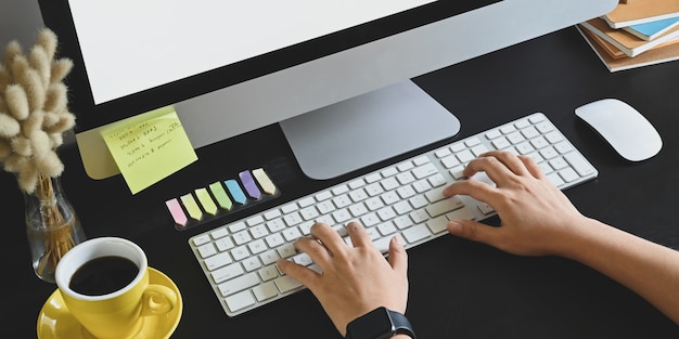 cropped image of hands typing on wireless keyboard that putting on black working desk with computer monitor, mouse, stack of books, coffee cup, wild grass in vase and post it.