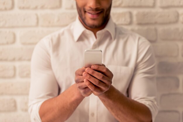 Cropped image of handsome Afro American businessman in classic shirt and tie using a smartphone and smiling against white brick wall