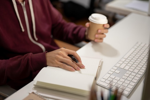 Photo cropped image of a man sitting in front of a computer taking notes in a book working in a room