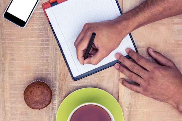 Cropped image of man writing on diary at table by coffee and cup cake