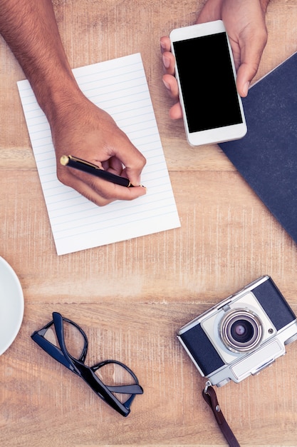 Immagine potata dell'uomo che scrive sul blocco note mentre tenendo lo smart phone dalla macchina fotografica e dagli occhiali sul tavolo