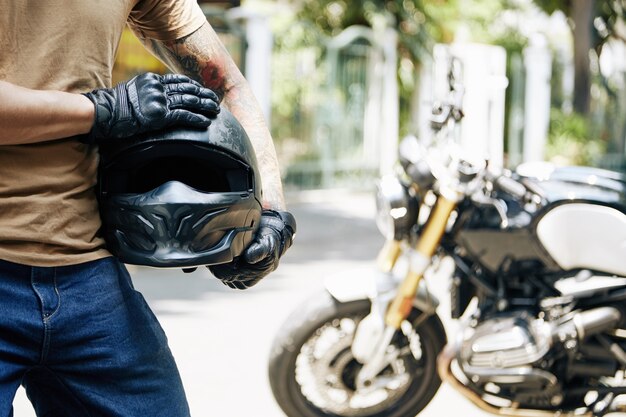Обрезанное изображение мотоциклиста в кожаных перчатках, держащего шлем, на заднем плане - его велосипед
