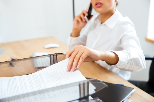 Фото Обрезанное изображение молодой деловой женщины разговаривает по телефону и принимает документы из офисного стола