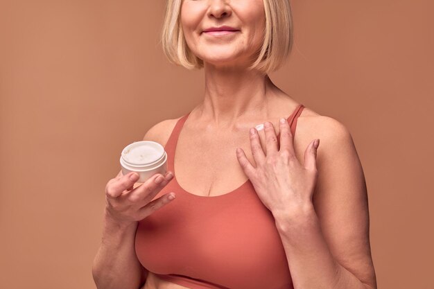 Cropped image of older woman in underwear holding cream in one hand and applying cream to collarbone with the otherBody care