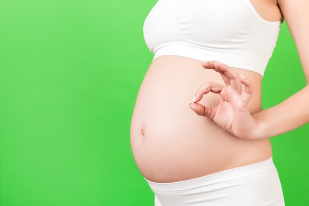 Обрезанное изображение позитивной беременной женщины в белом нижнем белье, показывающей нормальный жест против ее живота на зеленом фоне. Легкая и счастливая беременность. Скопируйте пространство.