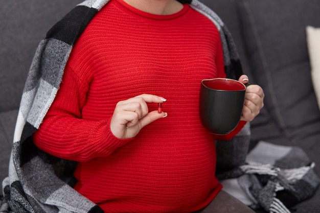 Cropped image of pregnant female takes drug, holds mug with tea, drinks medicine durning pregnancy, needs vitamins