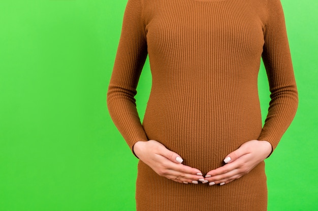 Cropped image of pregnant woman in brown dress holding her belly at green