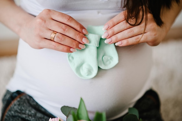 Cropped image pregnant woman holding socks for a baby on her belly stomach. Pregnant hugging tummy at home. Motherhood concept. Baby Shower.