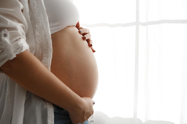 Cropped image pregnant woman standing near window in appartment strokes hugging the belly