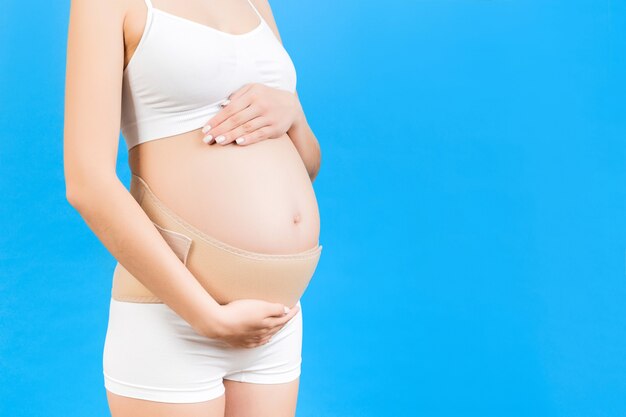 Cropped image of pregnant woman in underwear wearing maternity belt to reduce pain in the back at blue background with copy space. Orthopedic abdominal support belt concept.