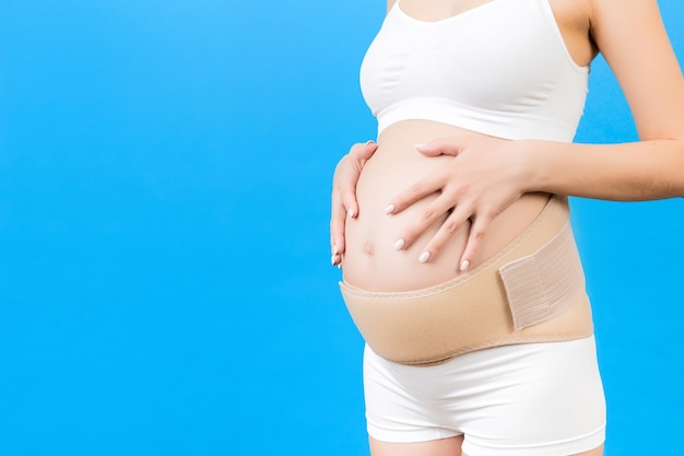 Cropped image of pregnant woman in underwear wearing maternity belt to reduce pain in the back at blue background with copy space. Orthopedic abdominal support belt concept.