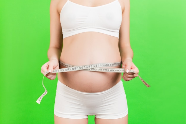 Cropped image of pregnant woman in white underwear measuring her abdomen to check baby development. Centimeter tape measure at green wall. Healthy pregnancy. Copy space