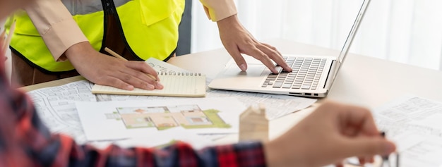 Cropped image of professional engineer team working on blueprint while his coworker working on laptop at meeting table with blueprint and wooden block scattered around Closeup Delineation