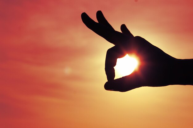 Cropped image of silhouette hand holding sun against sky during sunset