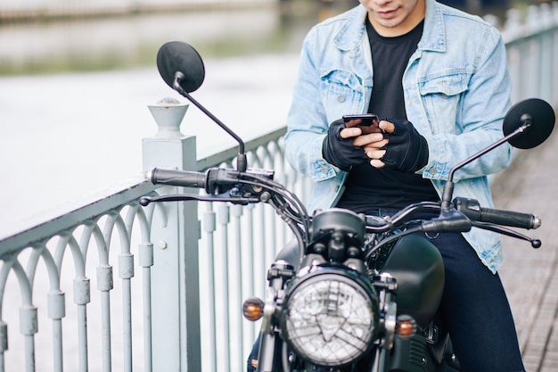 Photo cropped image of smiling biker in denim jacket and fingerless gloves sitting on motorcycle and texting friend or girlfriend