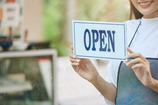 Cropped image of smiling cafe owner hanging open sign on glass door of cafe