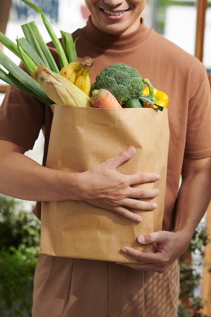 Cropped image of smiling young man holding big paper bag of fresh groceries from local market