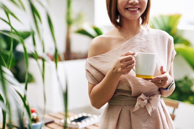 Обрезанное изображение улыбающейся молодой вьетнамской женщины, стоящей в кофейне с кружкой горячего напитка