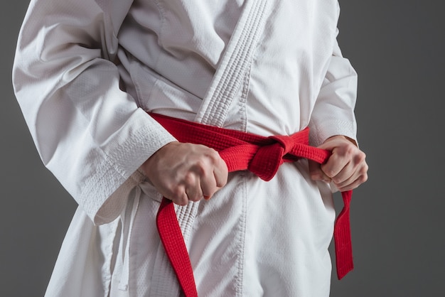 Cropped image of sportsman dressed in kimono practice in karate while tightening red belt isolated over grey background.