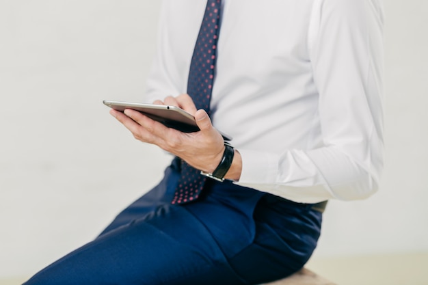 Cropped image of successful prosperous businessman in white shirt, black trousers, tie, holds modern tablet